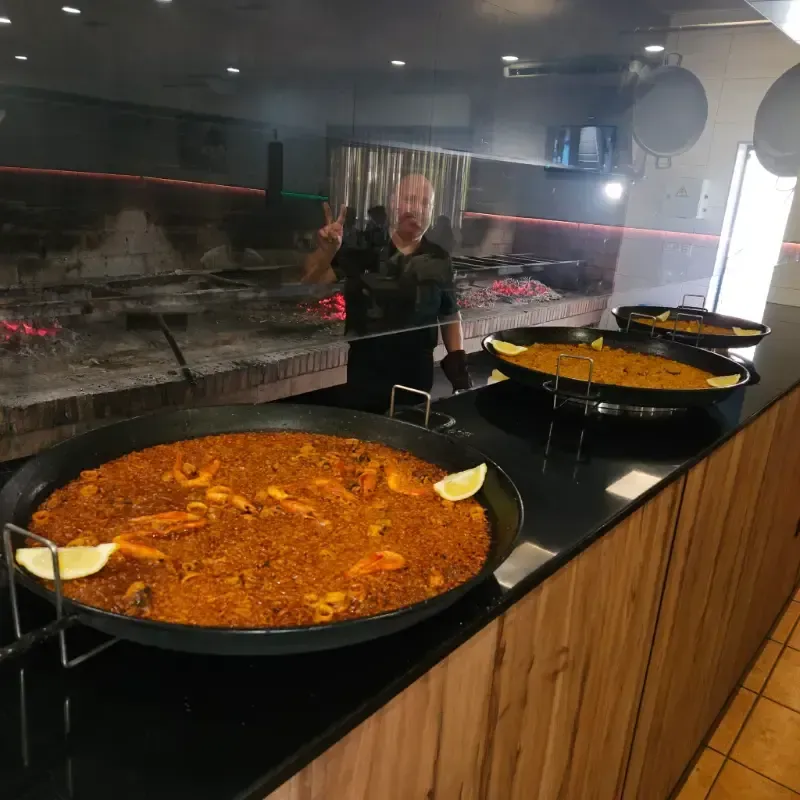 Un chef hace el signo de la paz detrás de un mostrador donde se exhiben grandes paelleras recién preparadas.