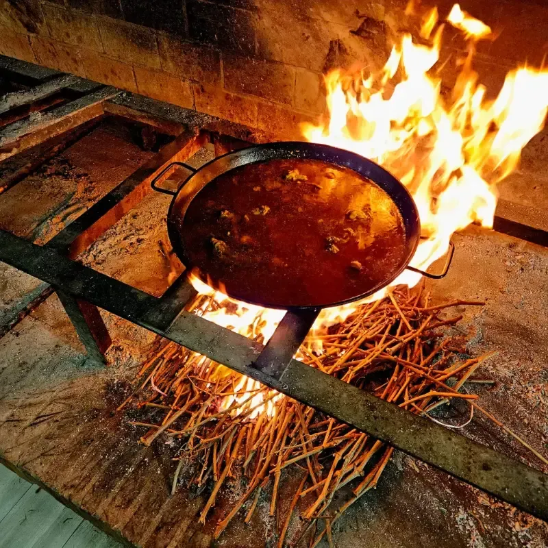 Una gran cacerola metálica llena de un guiso rojizo burbujeante reposa sobre un fuego de leña abierto
