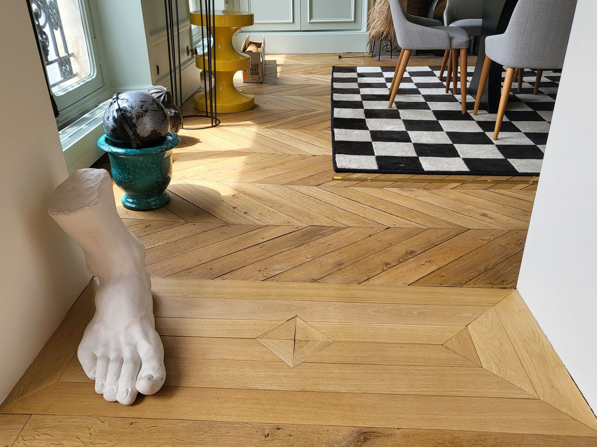 Une pièce avec parquet, une sculpture blanche représentant un pied et un tapis à carreaux près d'une table à manger.