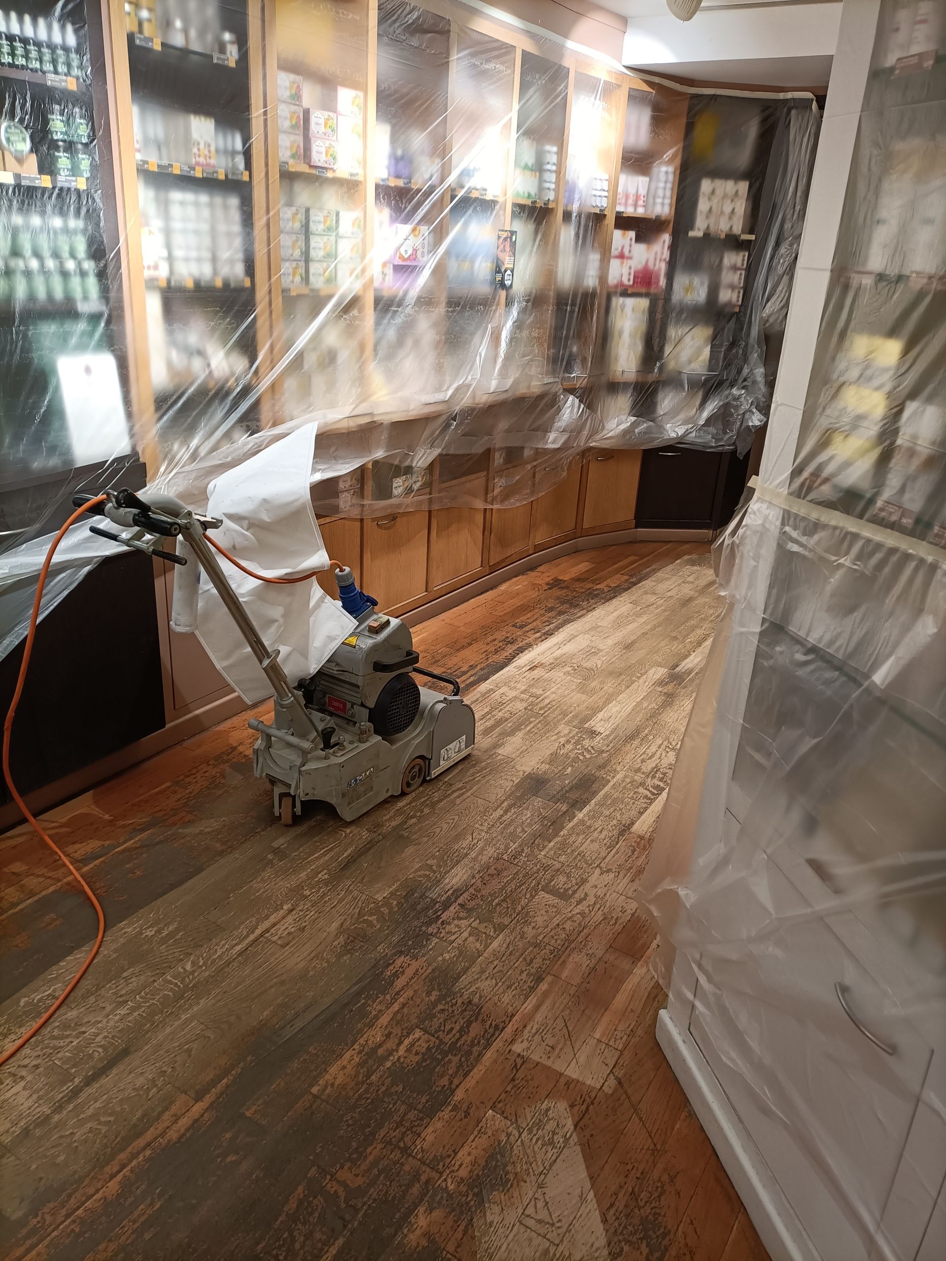 Ponceuse à parquet dans un magasin, sol en bois partiellement poncé, étagères recouvertes de plastique de protection.