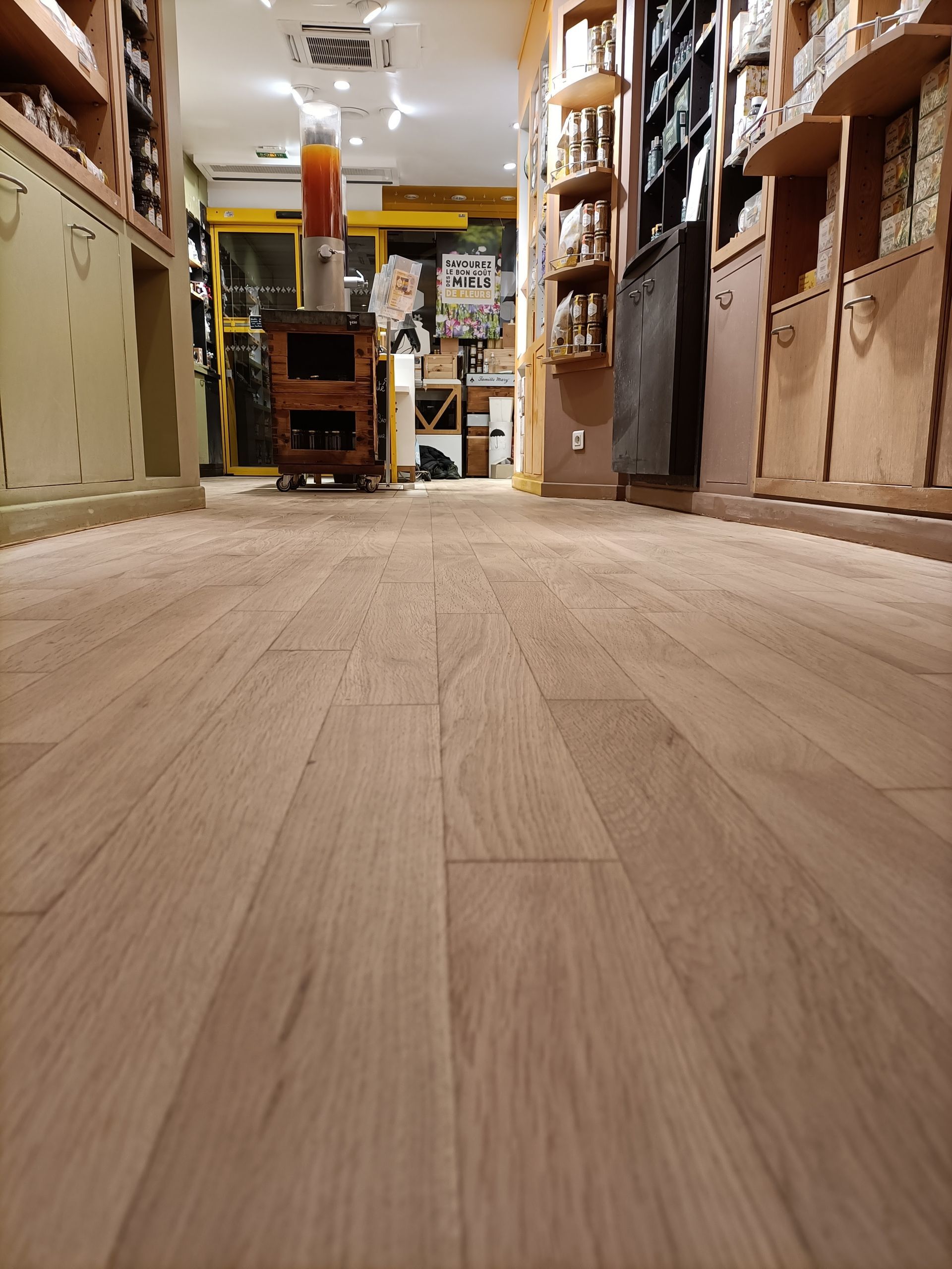 Allée d'un magasin avec un sol en bois, bordée d'étagères remplies de marchandises, éclairée par des lampes suspendues.