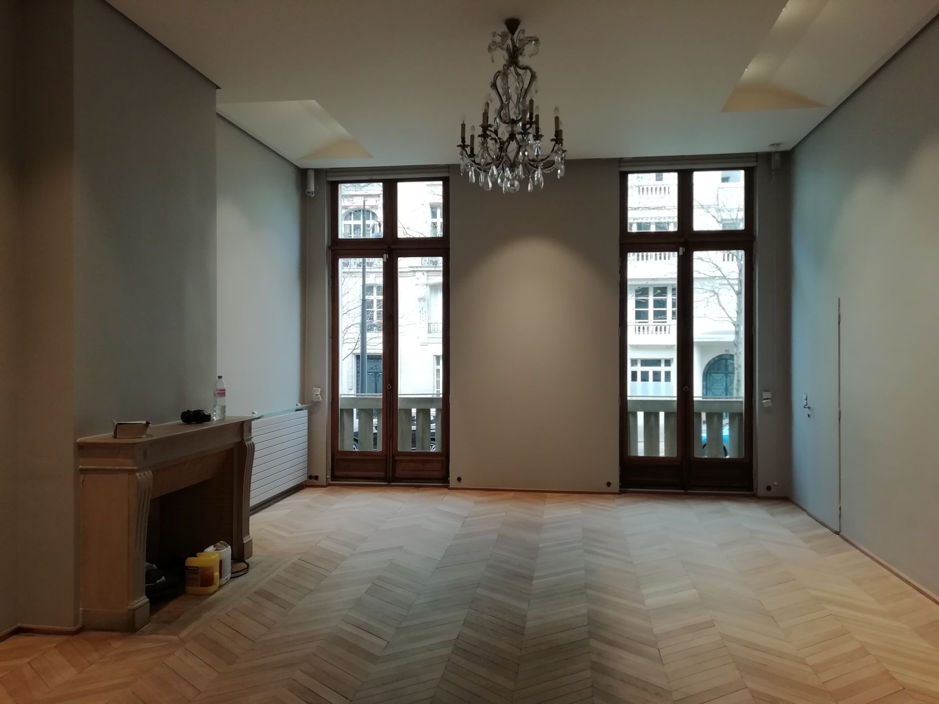 Pièce vide avec parquet, cheminée, deux grandes fenêtres et un lustre.