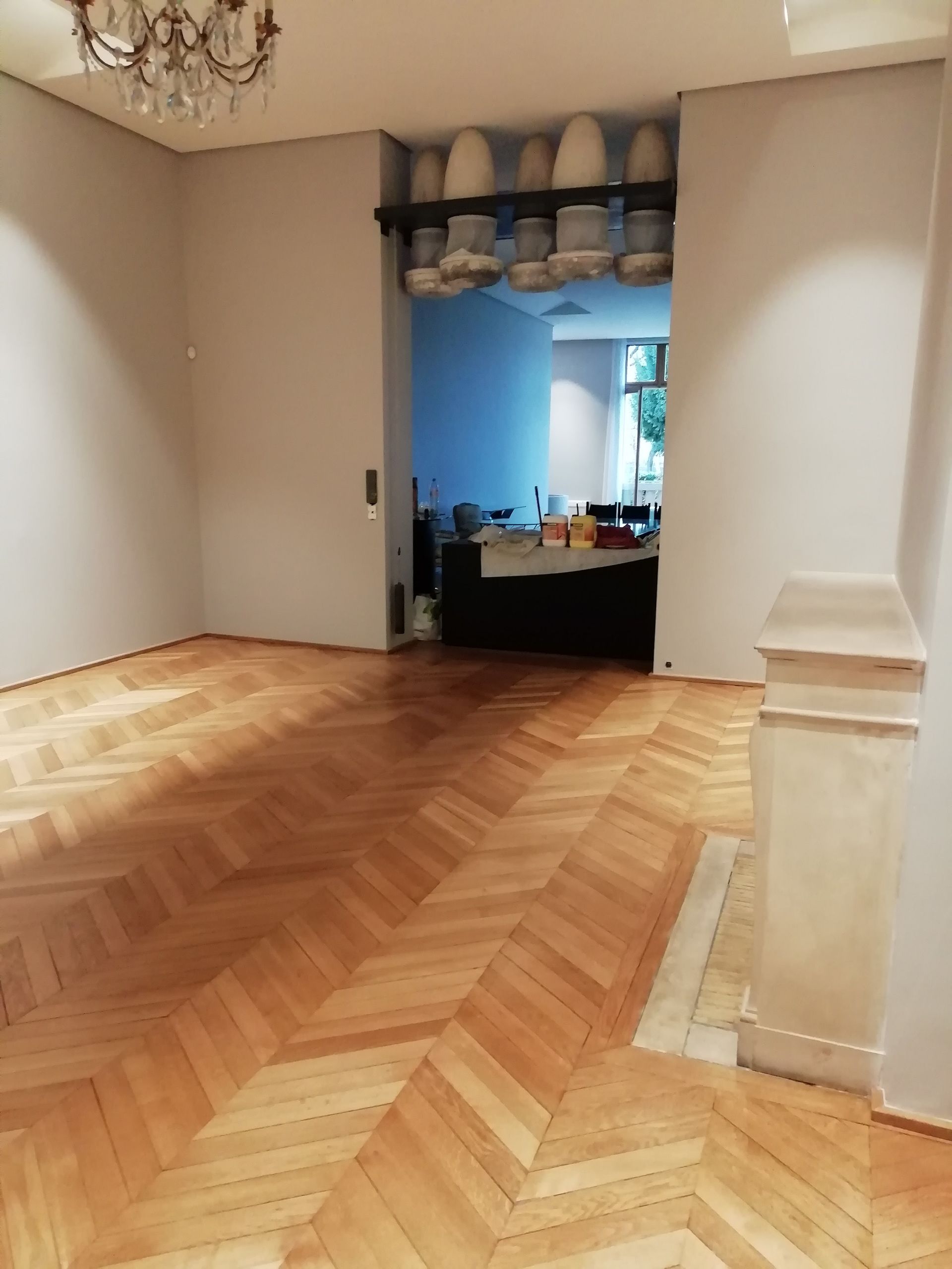 Pièce vide avec parquet à chevrons, porte donnant sur une autre pièce, murs blancs et pilier décoratif.