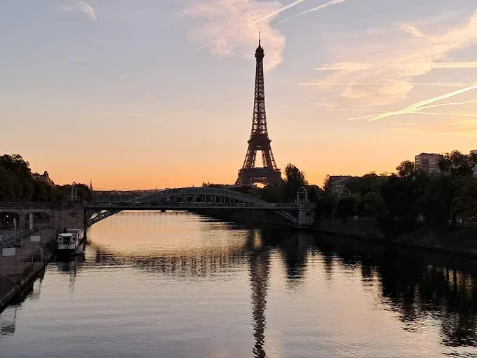 La Torre Eiffel al atardecer reflejada en el Sena con un puente en primer plano.