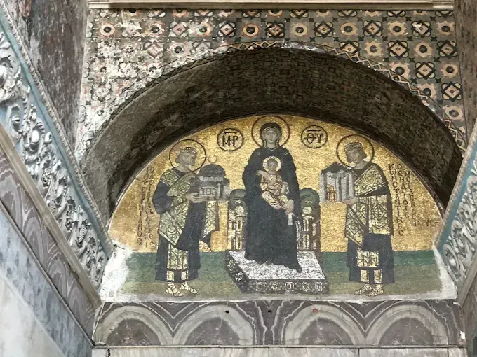 Mosaico bizantino de tres figuras con aureolas bajo una hornacina arqueada, con fondo de piedra dorada y decorada.