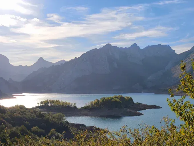 Lago de montaña al amanecer con aguas azules tranquilas, picos oscuros y una pequeña isla verde.