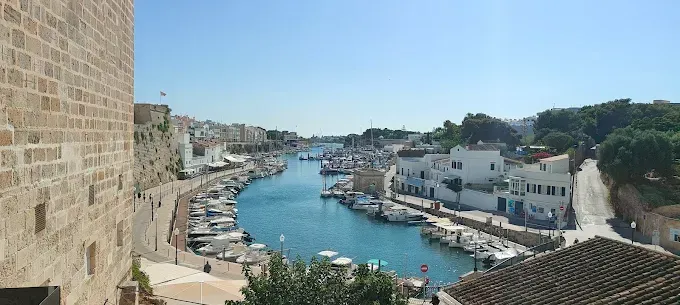 Pueblo portuario con barcos amarrados a lo largo de un canal azul entre edificios blancos y muros de piedra.