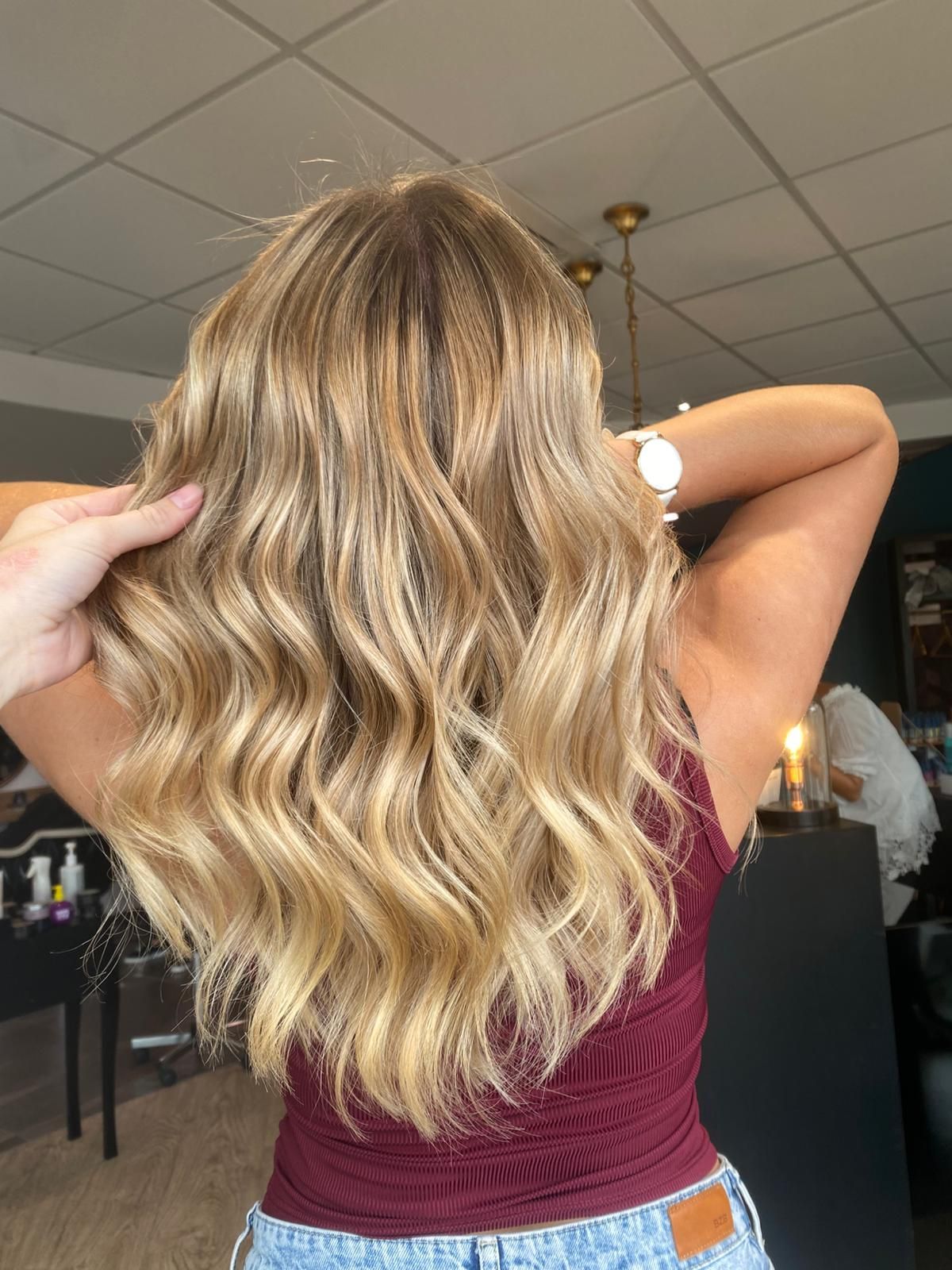 Femme aux cheveux ondulés blonds et châtain clair, portant un haut bordeaux, dans un salon de coiffure.