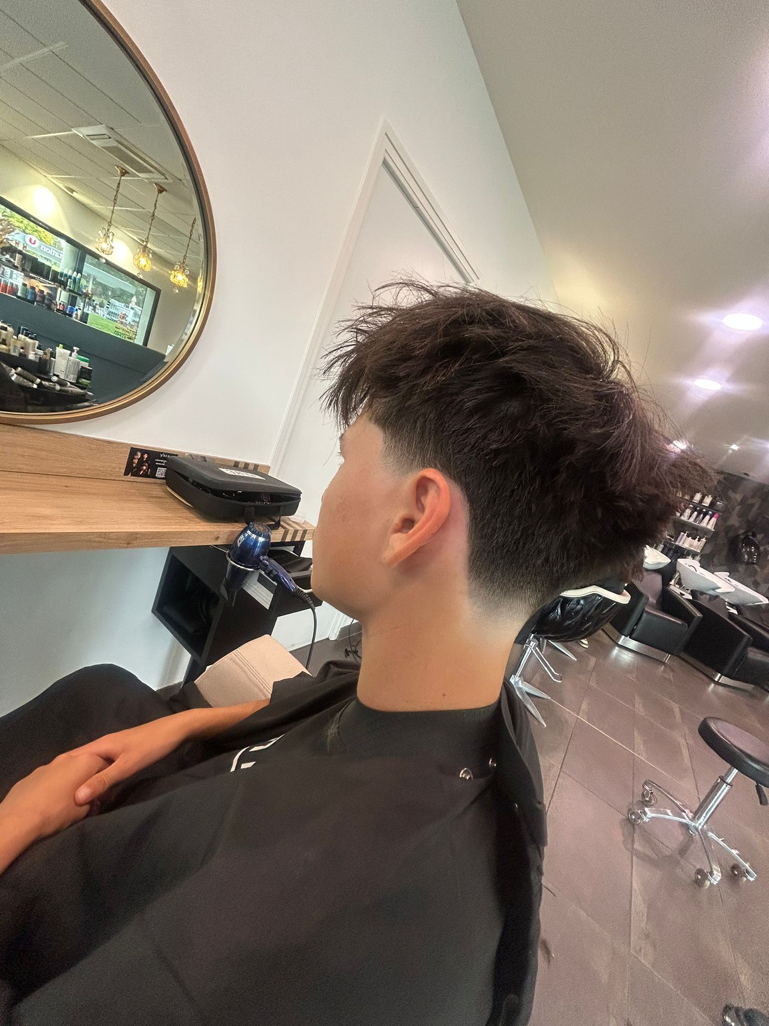 Personne aux cheveux bouclés, coupe dégradée, assise dans un salon de coiffure, portant une cape de barbier.