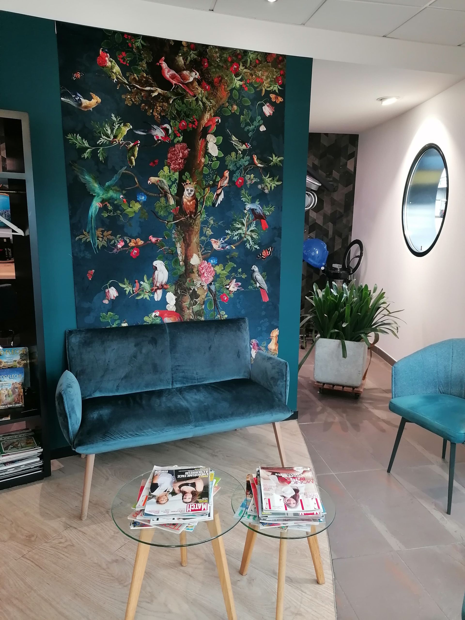 Canapé et fauteuils bleu canard dans une salle d'attente avec une tapisserie florale, un miroir rond et des tables basses en verre.