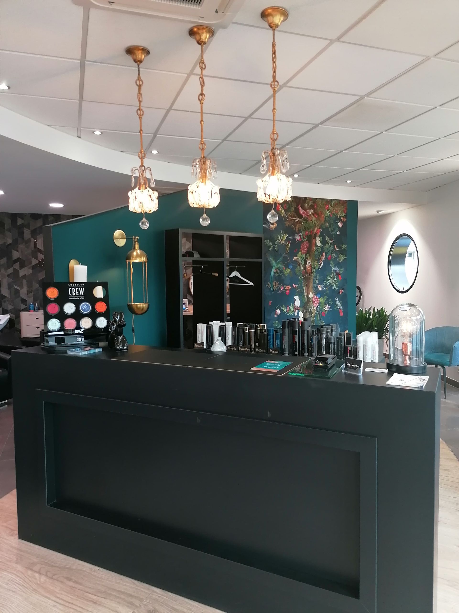 Un espace d'accueil de salon avec un comptoir noir, un mur turquoise et des suspensions lumineuses dorées.