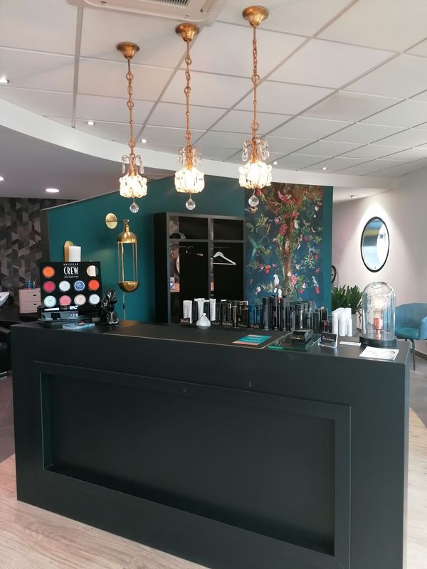 Un espace d'accueil de salon avec un comptoir noir, un mur turquoise et des suspensions lumineuses dorées.