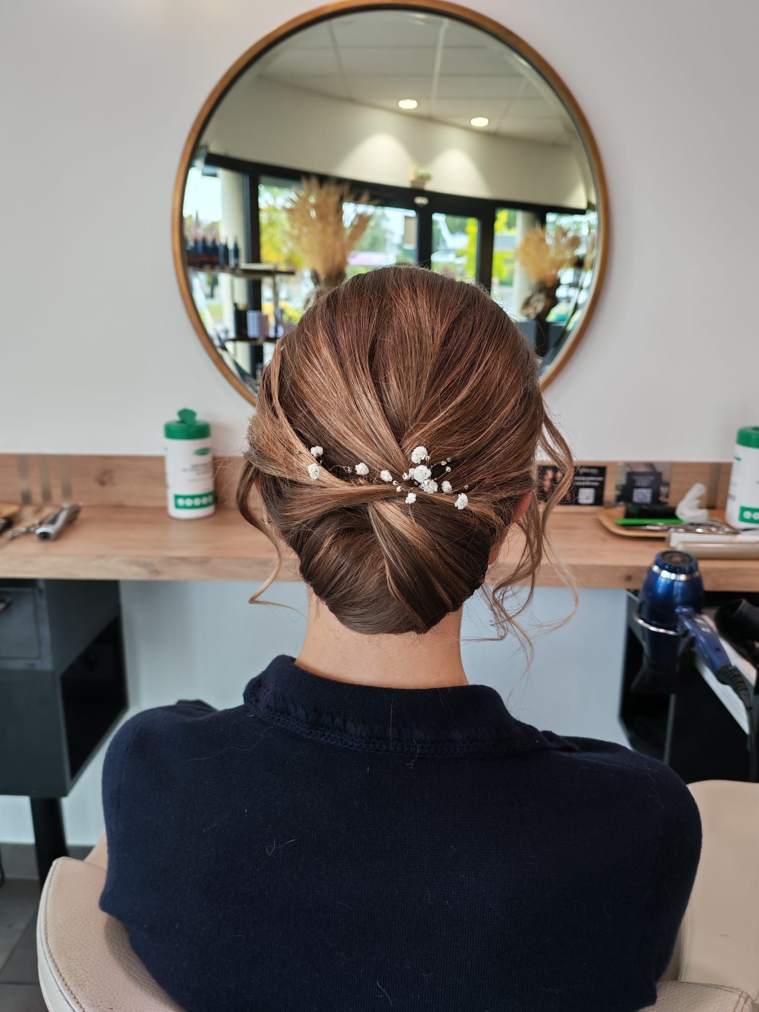 Femme aux cheveux bruns coiffés en chignon, devant un miroir dans un salon de coiffure. Ses cheveux sont ornés d'épingles à cheveux en perles.