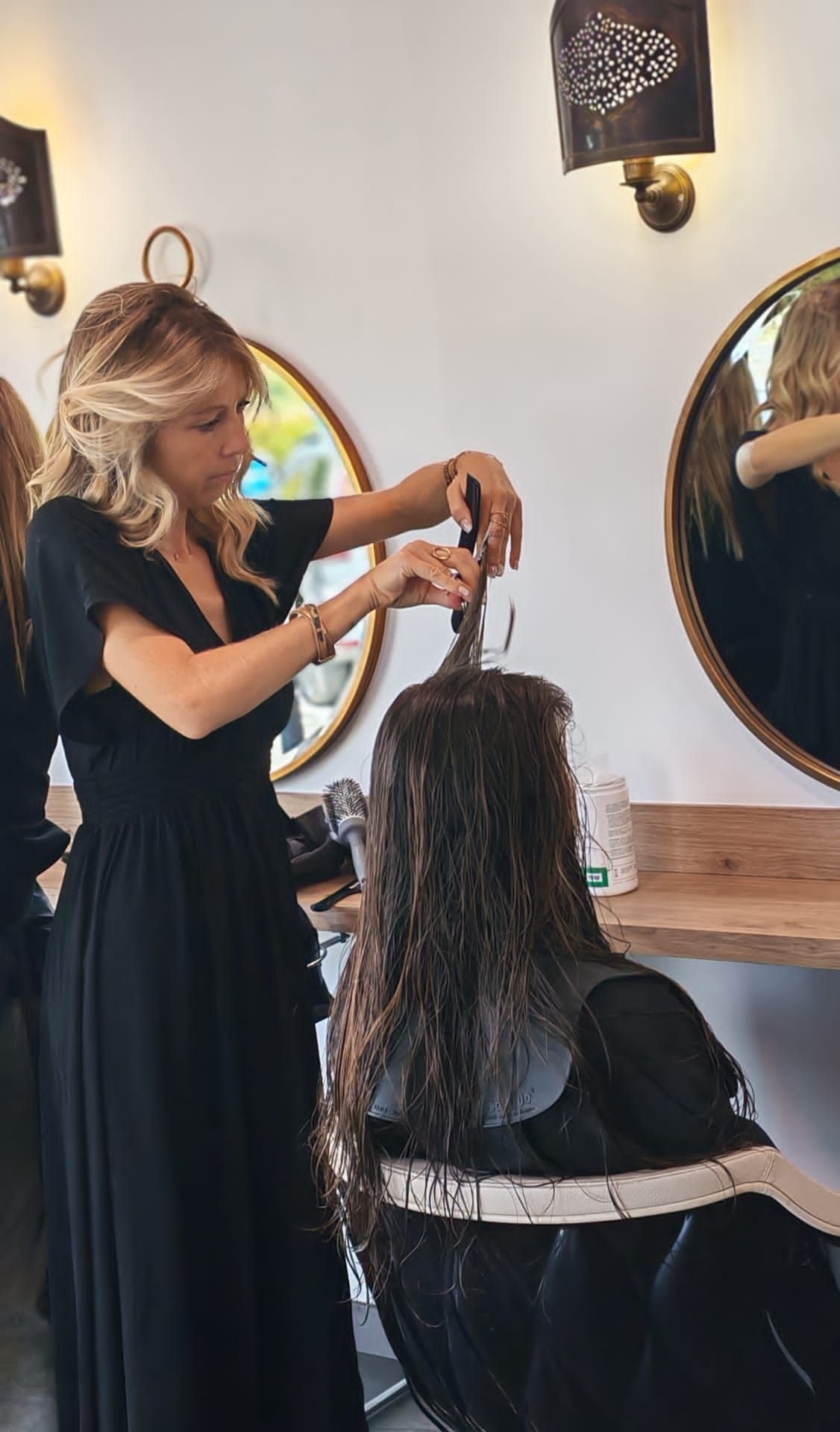 Dans un salon de coiffure, une coiffeuse coupe les cheveux mouillés d'une cliente, près d'un miroir. Toutes deux sont vêtues de noir.