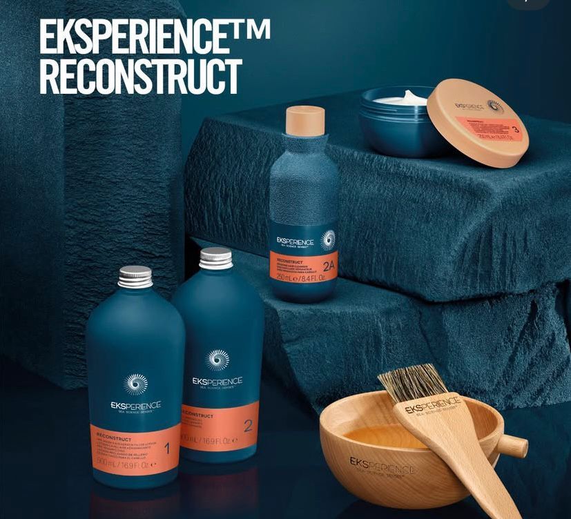 Produits capillaires EKSperience Reconstruct : flacons, pots, bol et brosse sur une surface texturée bleue.