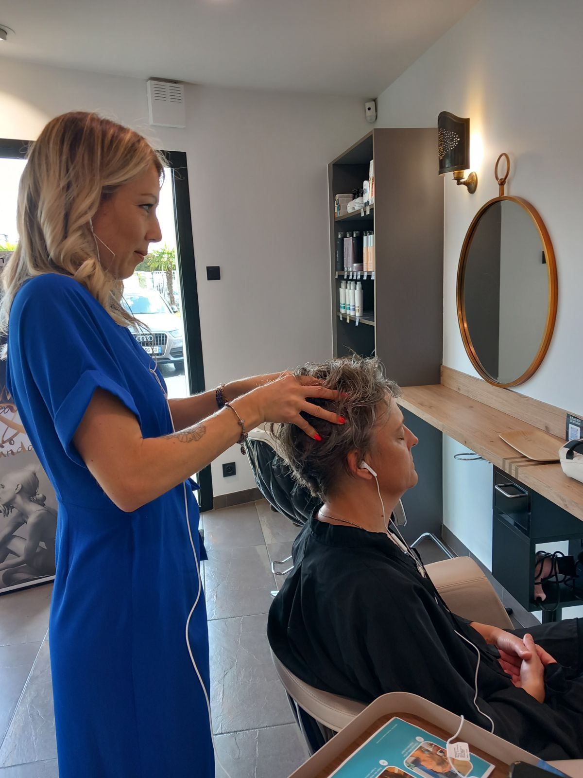 Une femme en robe bleue masse la tête d'une autre femme dans un salon de coiffure. Miroir et étagères en arrière-plan.