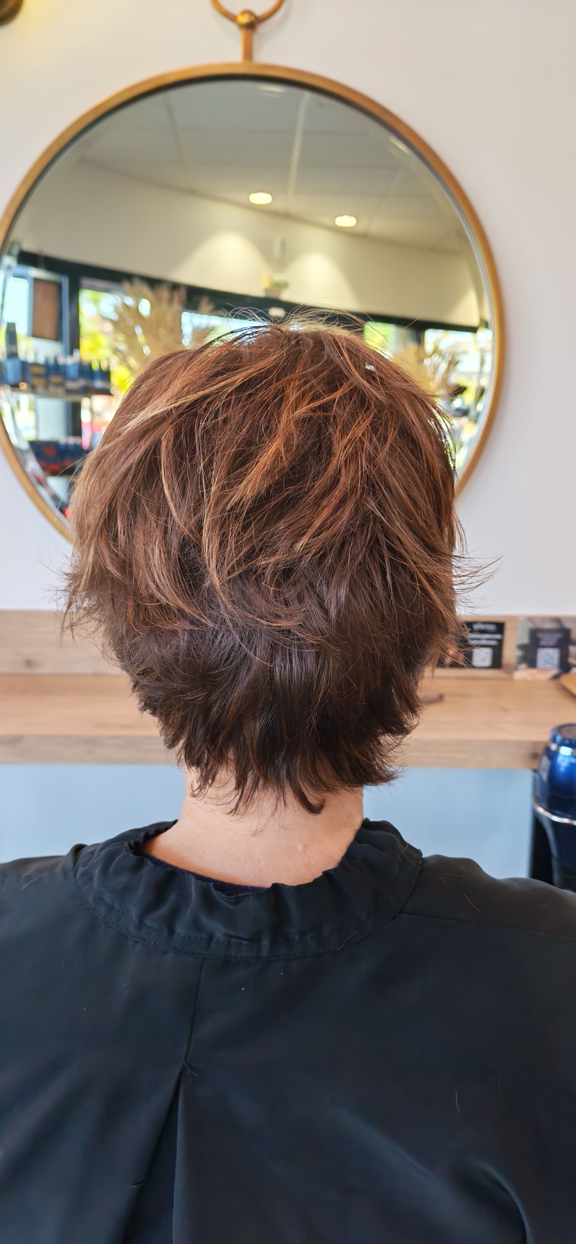 Vue de dos d'une personne aux cheveux courts, bouclés et bruns, devant un miroir rond, dans un salon de coiffure.