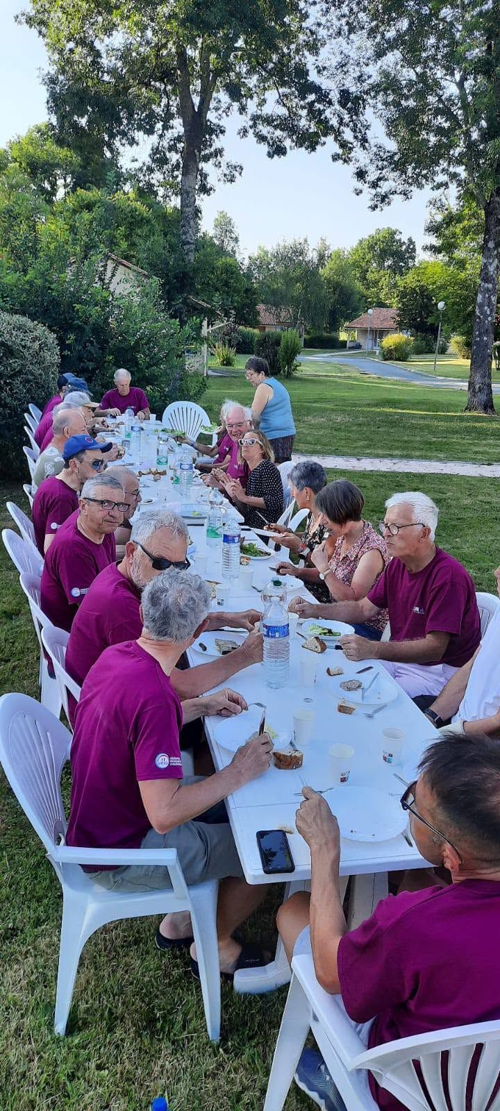 Des personnes assises à une longue table en plein air, sous des arbres, partagent un repas ou une activité dans un jardin.