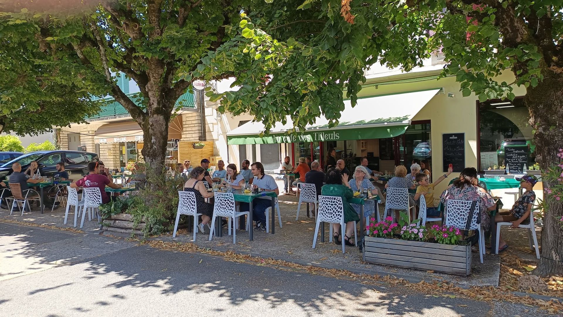 Des personnes assises à des tables sous des arbres à l'extérieur d'un bâtiment, rassemblées dans un café ombragé en plein air.