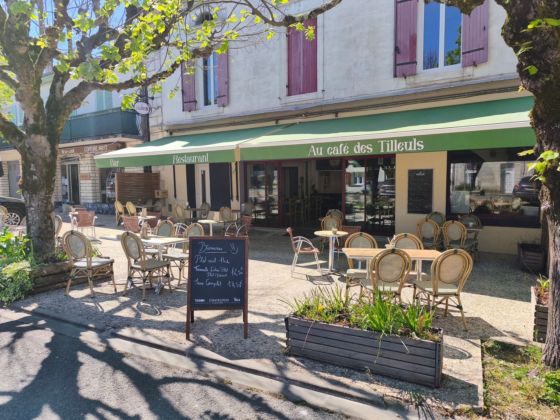 Terrasse de café sous un auvent vert, avec tables, chaises et menu écrit à la craie sur un tableau noir, dans une rue ensoleillée.