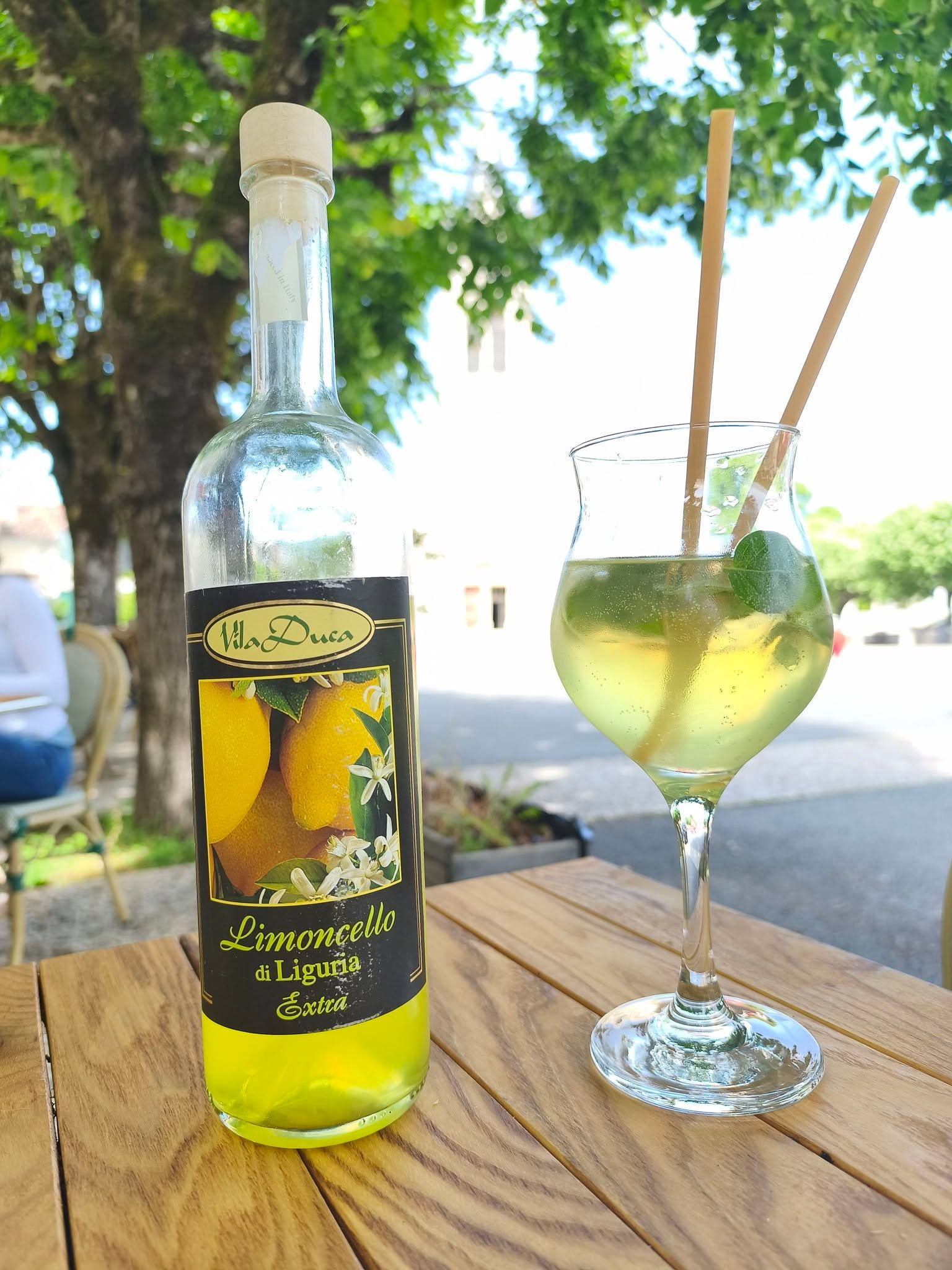 Bouteille de liqueur jaune sur une table en bois à l'extérieur, avec un verre à pied et une paille en arrière-plan.