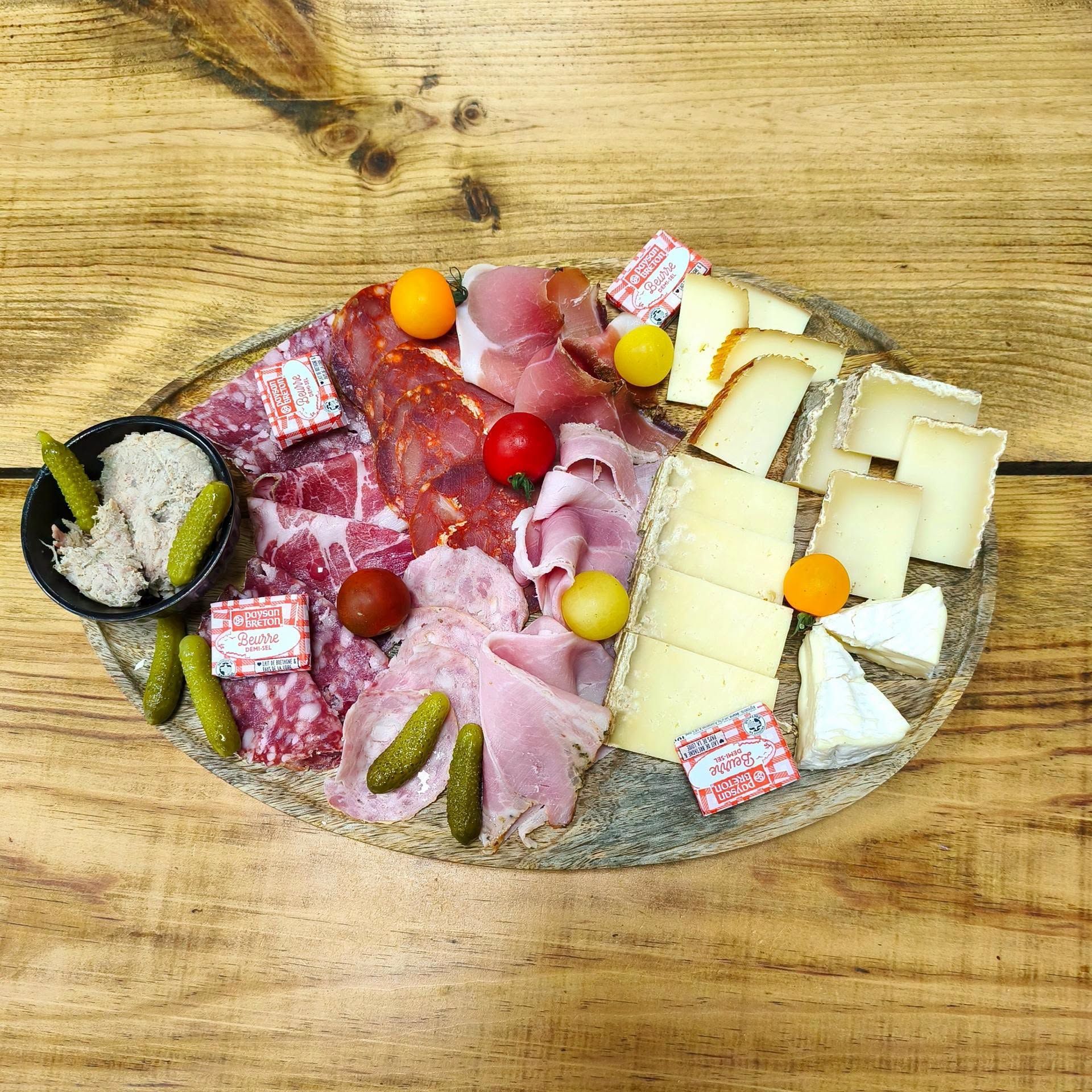 Planche de charcuterie avec viandes tranchées, fromages, cornichons et tomates cerises sur une table en bois.