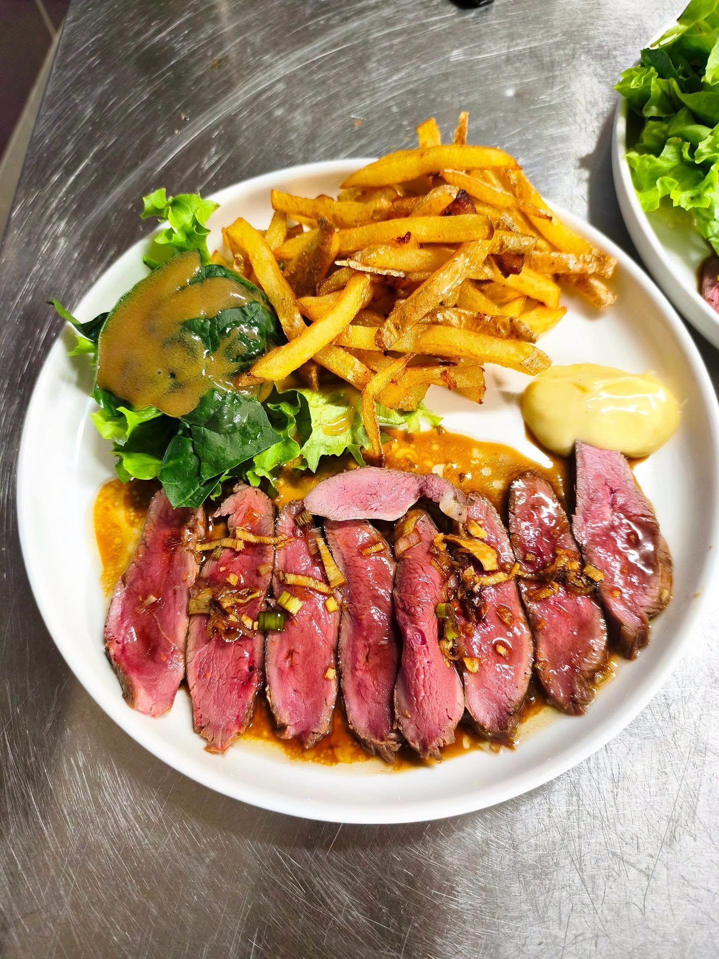 Steak tranché servi avec des frites, de la salade et de la sauce sur une assiette blanche, sur une table en acier inoxydable.