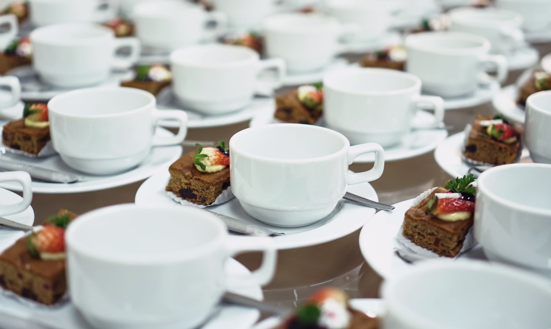 Des rangées de tasses et de soucoupes blanches sur les tables, avec des petits desserts présentés à l'assiette lors d'un événement formel.