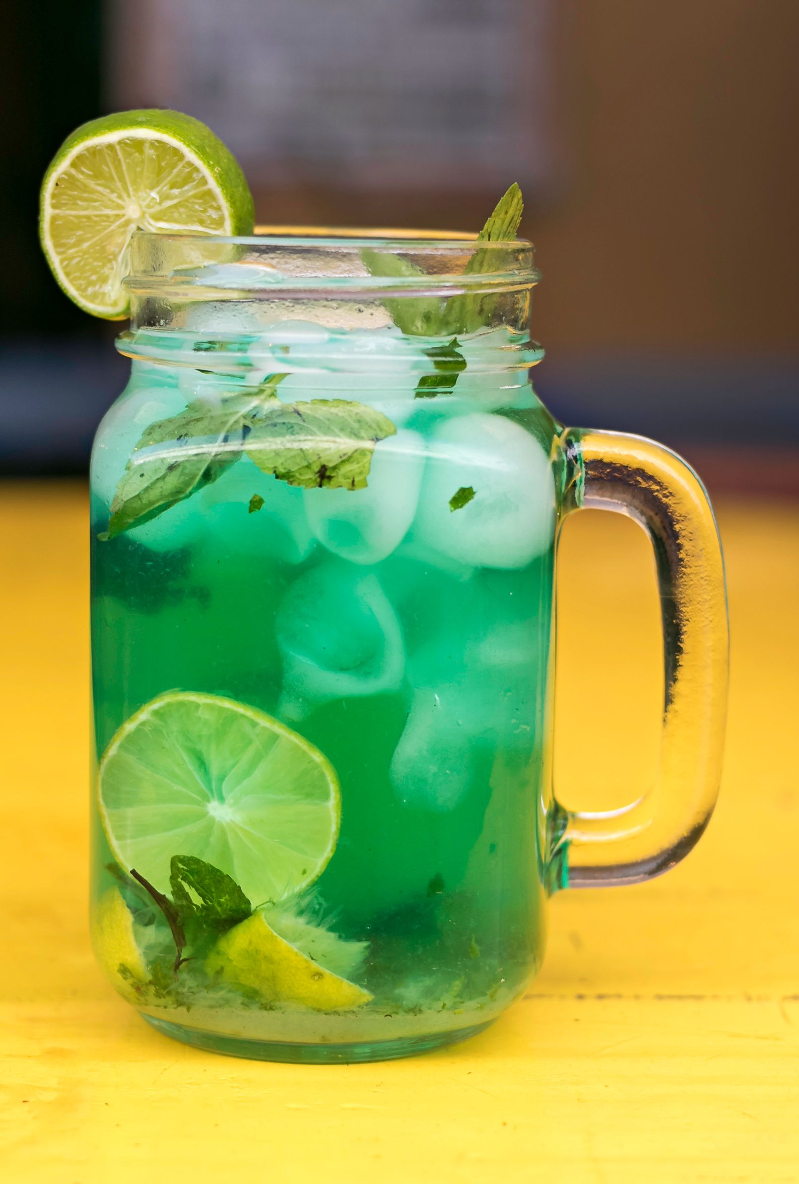 Boisson glacée verte dans un bocal en verre avec des tranches de citron vert et une paille sur une table jaune.
