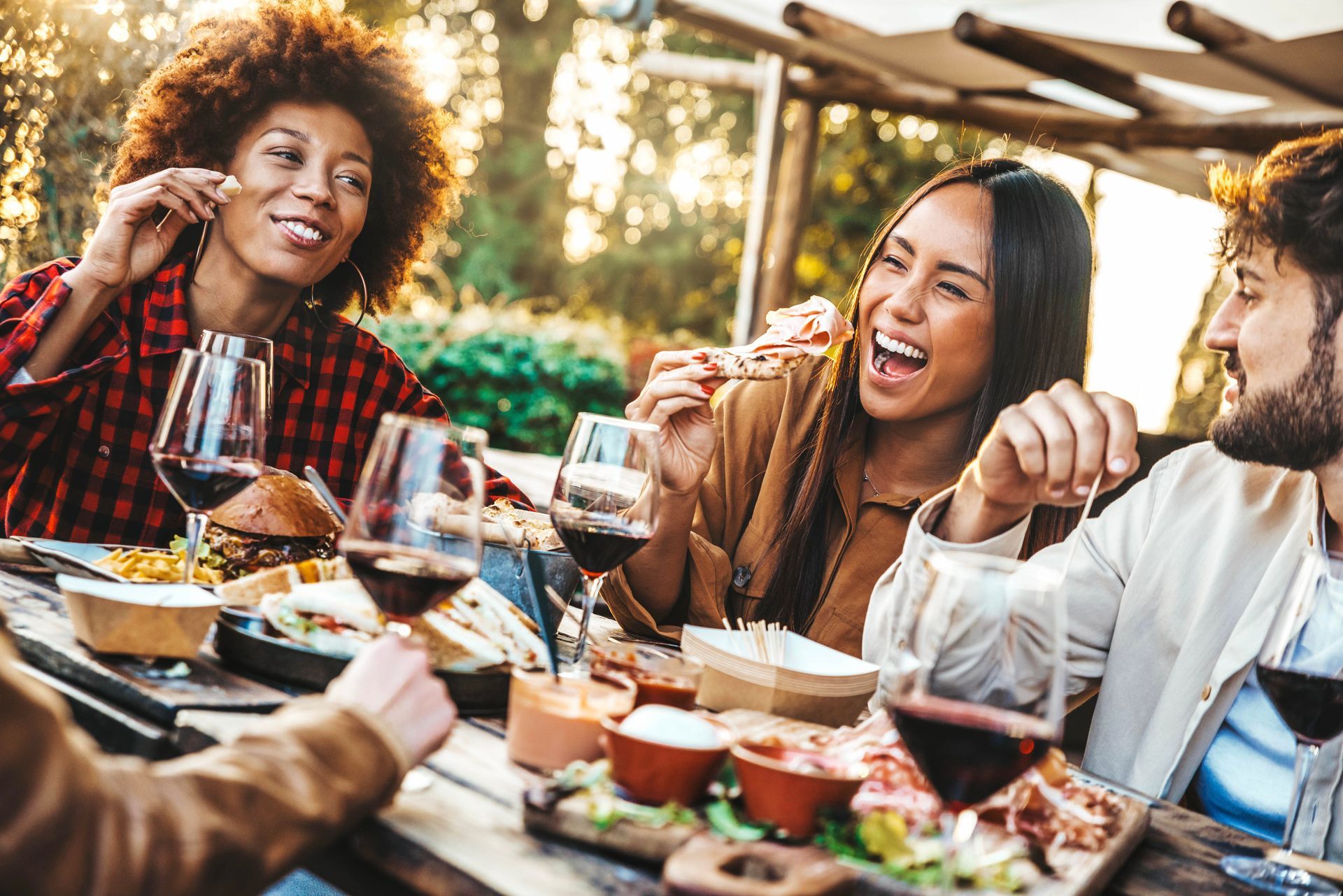 Des amis rient et partagent un repas et des boissons à une table d'extérieur sous un soleil radieux.