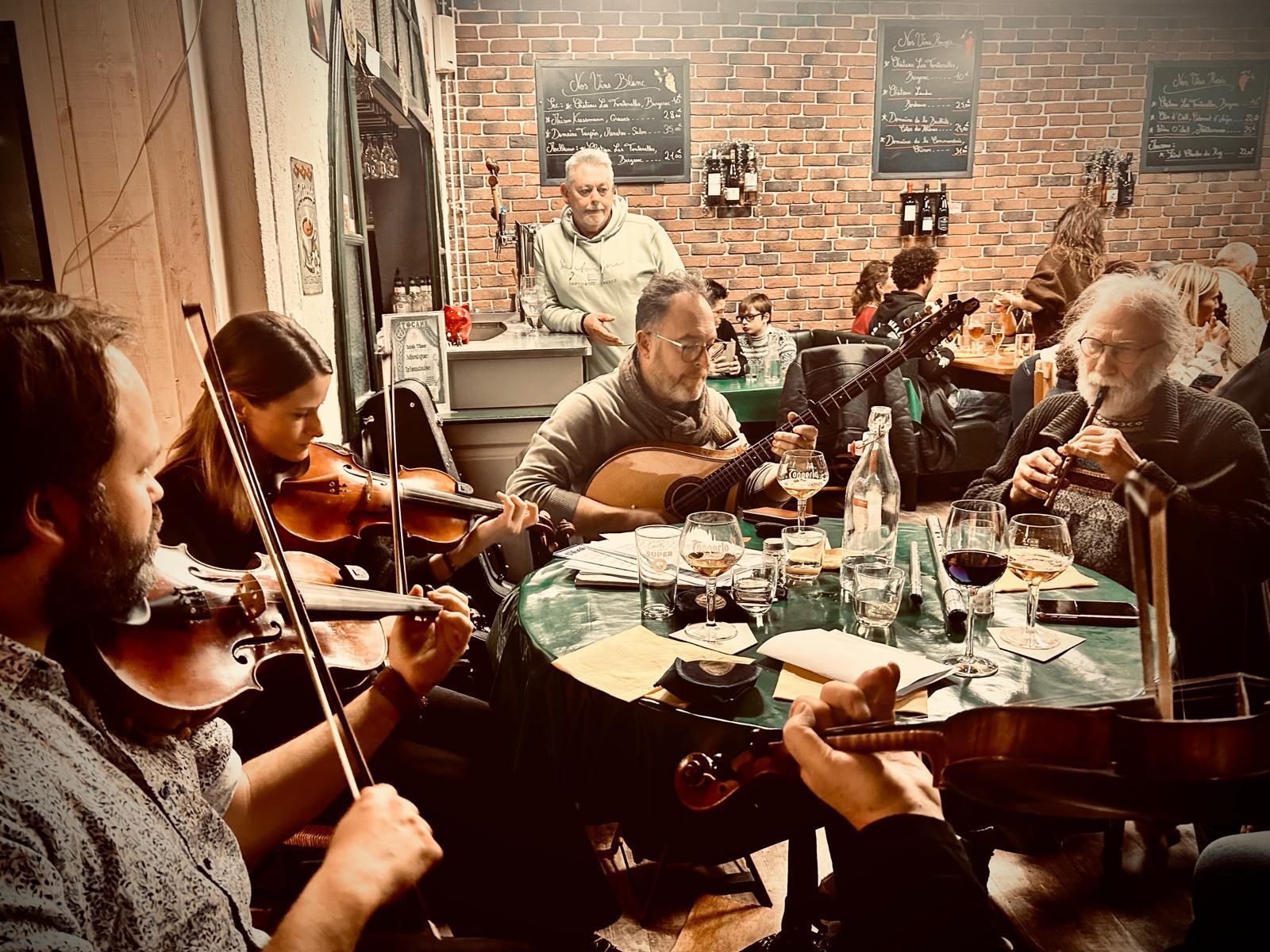 Des musiciens jouent du violon et du violoncelle autour d'une table dans une pièce chaleureuse aux murs de briques, avec des plats et des boissons à proximité.