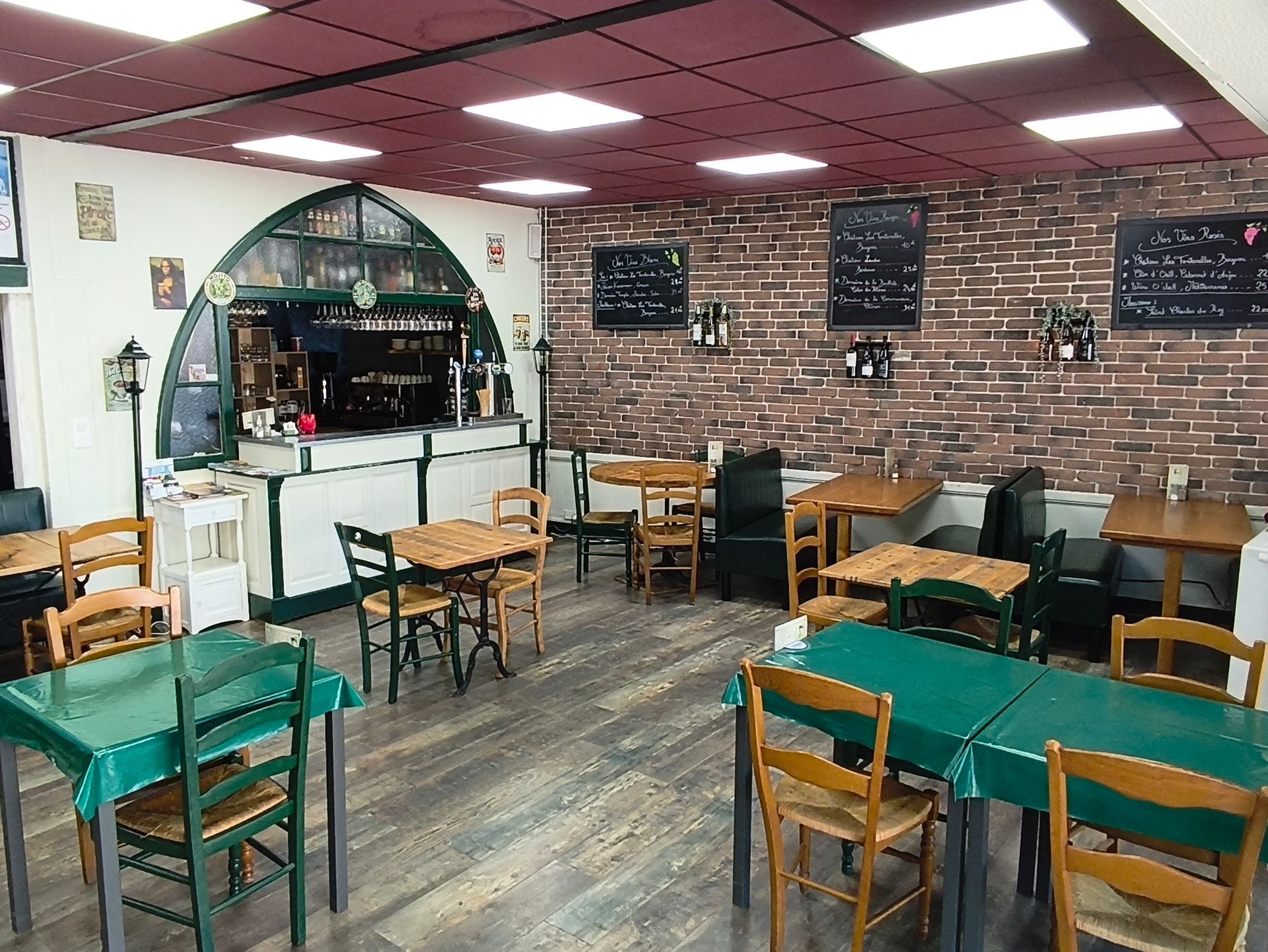 Café chaleureux aux murs de briques, avec nappes vertes, chaises en bois et un comptoir de service au fond.