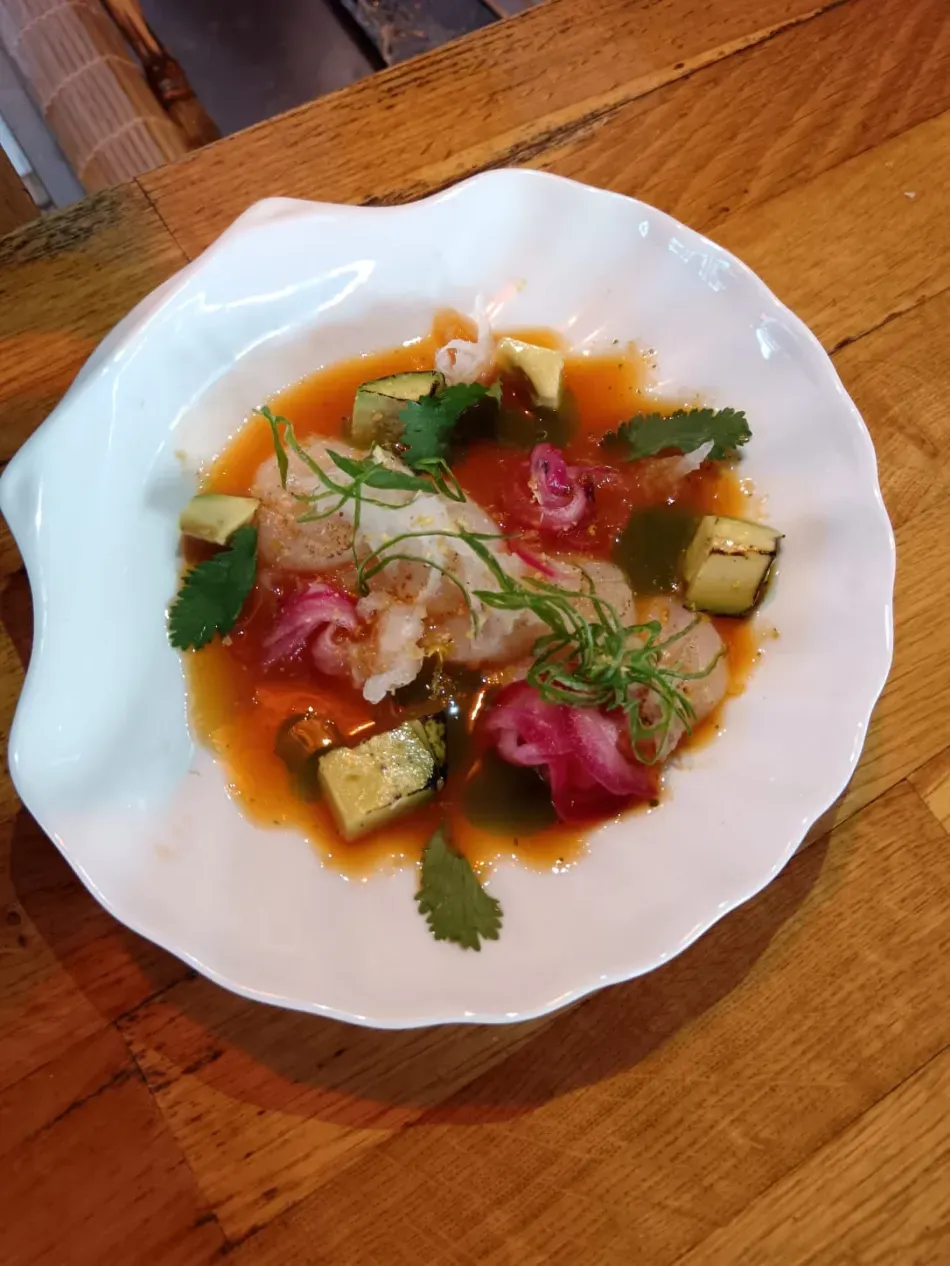 Plato gourmet emplatado con salsa verde, guarnición roja y verduras picadas en un plato blanco sobre una mesa de madera.