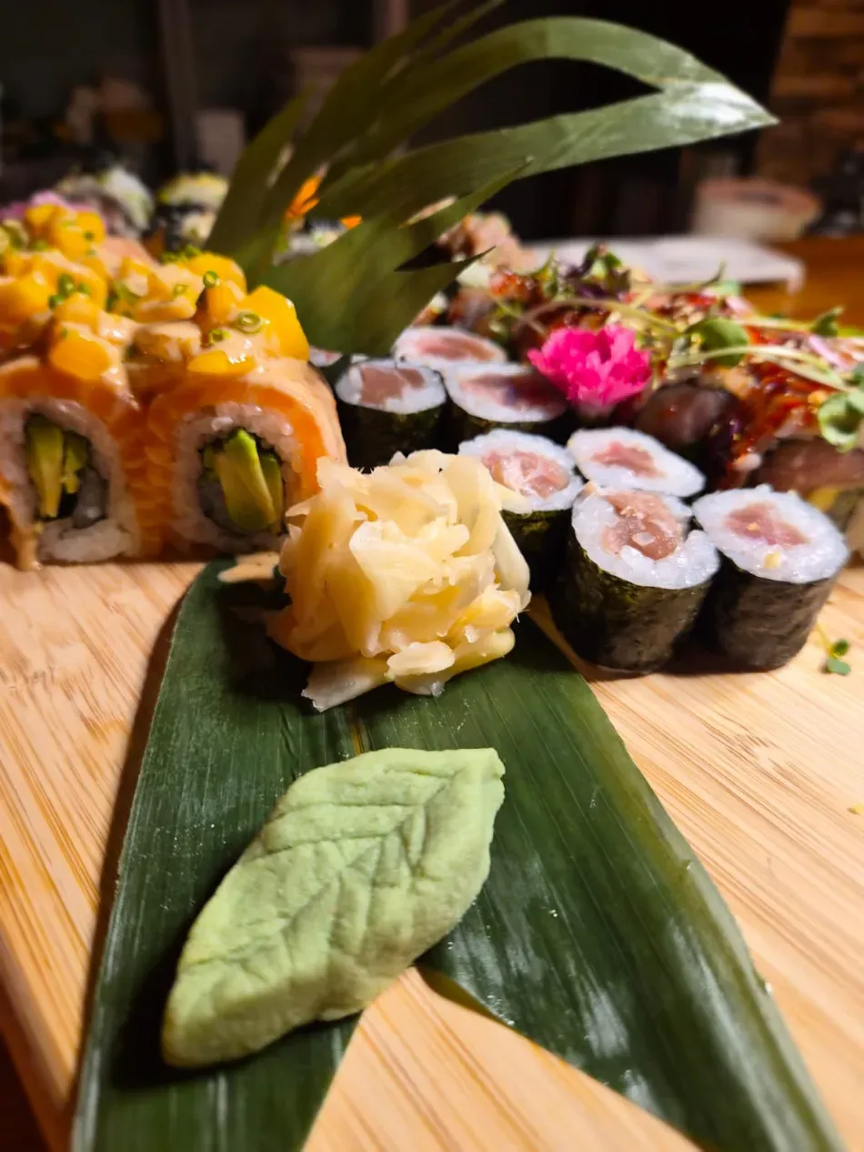 Rollos de sushi y guarniciones dispuestos sobre una tabla de madera decorada con hojas verdes.