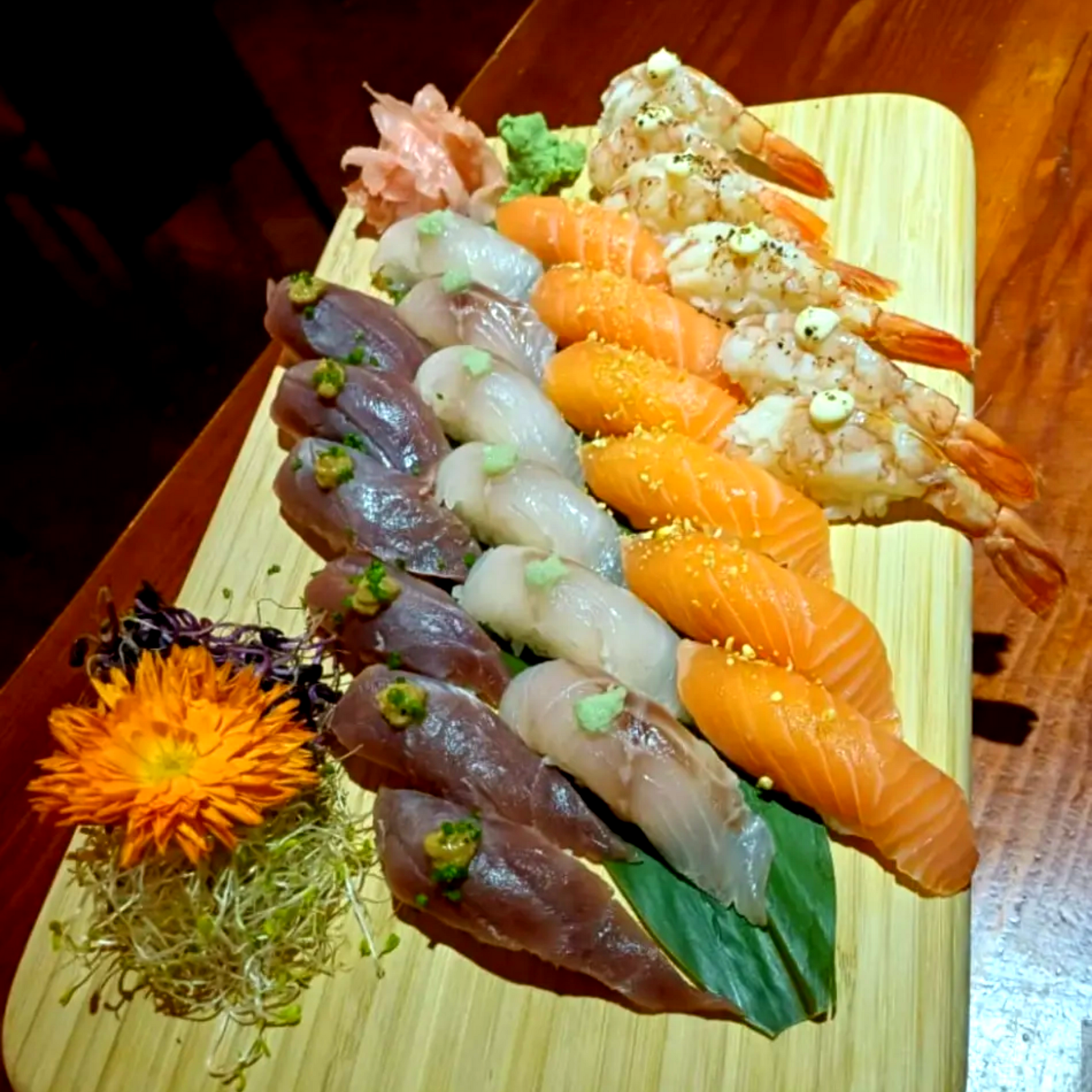Surtido de nigiri de sushi dispuesto en una tabla de madera con una flor de azahar como adorno.