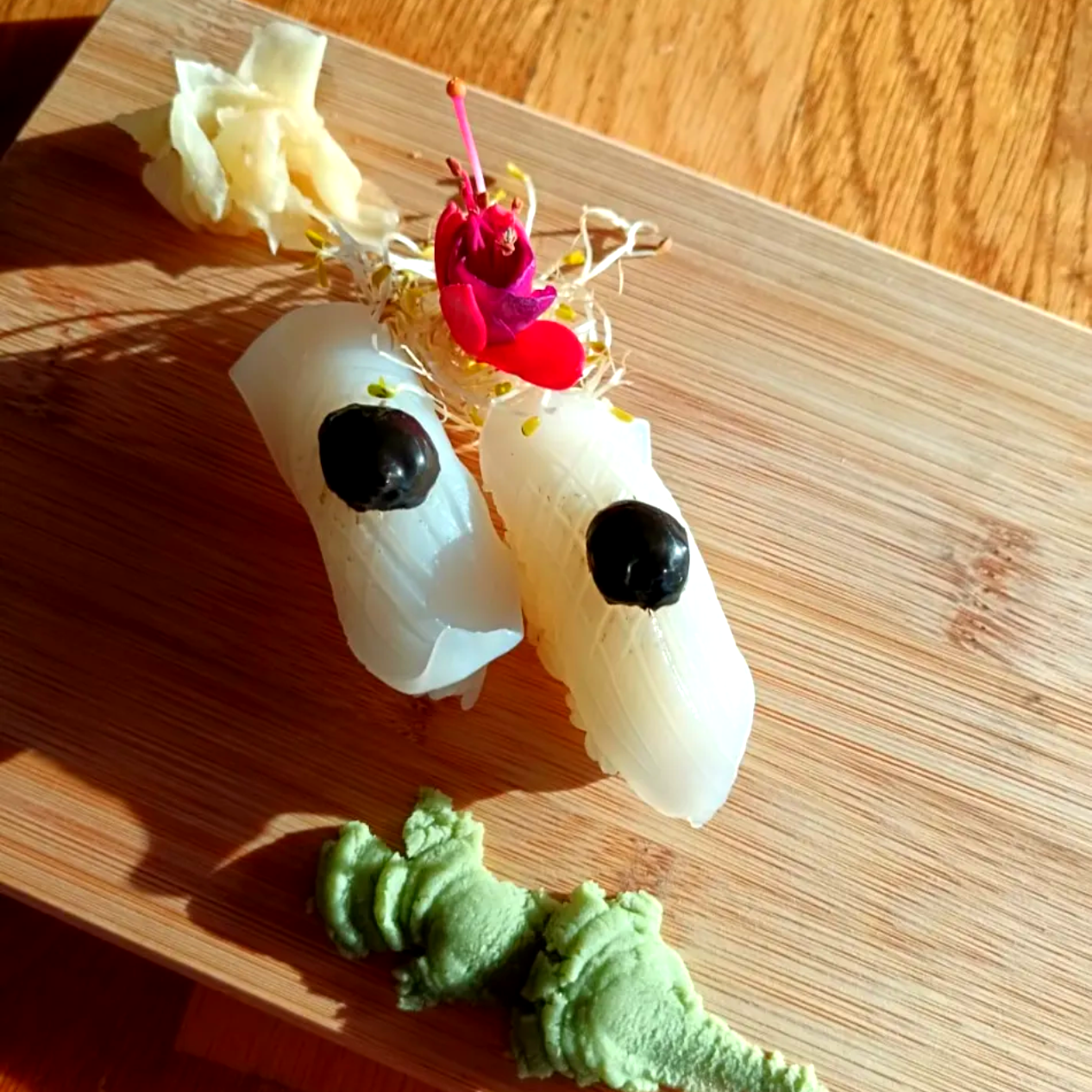 Dos pequeñas figuras blancas parecidas a granos de arroz con ojos negros sobre una tabla de madera, adornadas con detalles verdes y rosas.