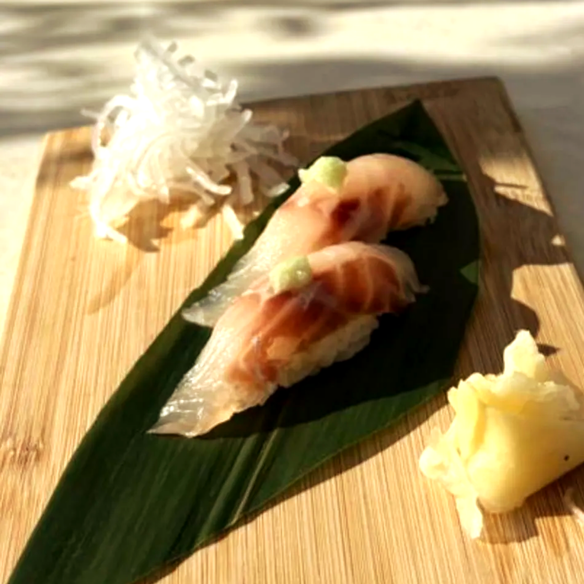 Dos piezas de sushi sobre una hoja verde con jengibre encurtido y wasabi en un plato de madera.