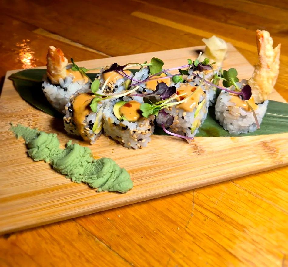 Rollos de sushi sobre una tabla de madera, cubiertos con hierbas y salsas, con trozos de tempura en los extremos.