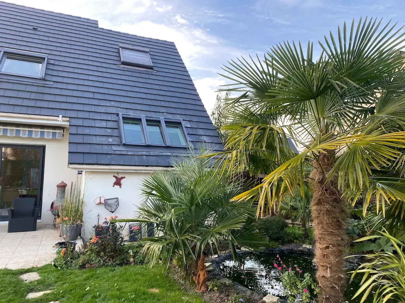 Maison au toit bleu et aux murs blancs, jouxtant un jardin luxuriant planté de palmiers.