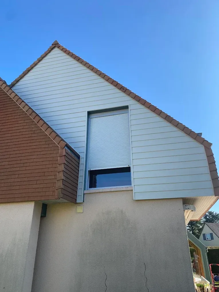 Extérieur d'une maison d'habitation avec un bardage blanc, un mur latéral en briques et une fenêtre à l'étage partiellement recouverte d'un store.