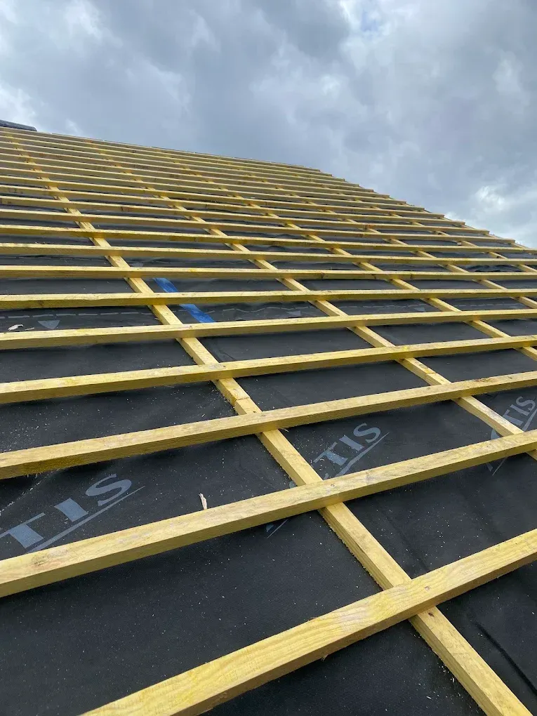 Lattes de toit en bois jaune sur une membrane de toiture noire, le tout sur fond de ciel nuageux.