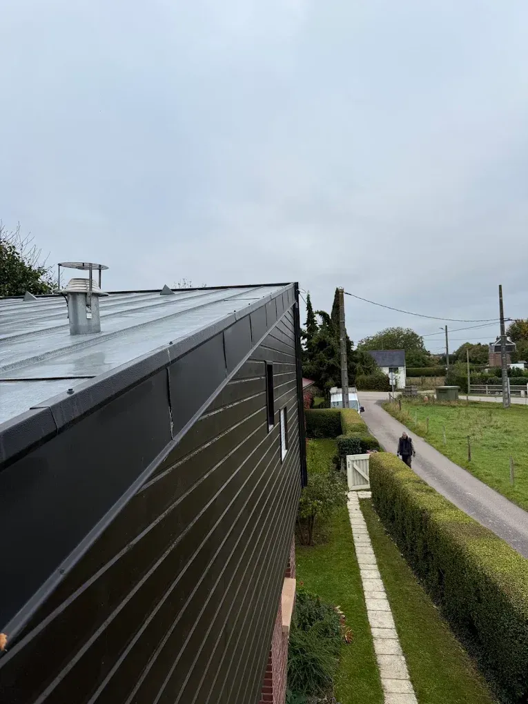 Vue de rue à côté d'un bâtiment noir au toit en pente, d'une haie, d'un trottoir et d'un ciel nuageux.