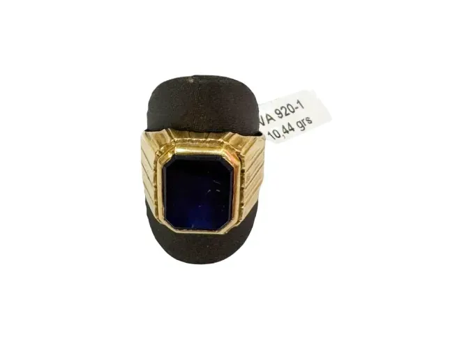 Anillo de oro con una piedra preciosa cuadrada de color azul oscuro sobre un soporte negro, con una etiqueta.