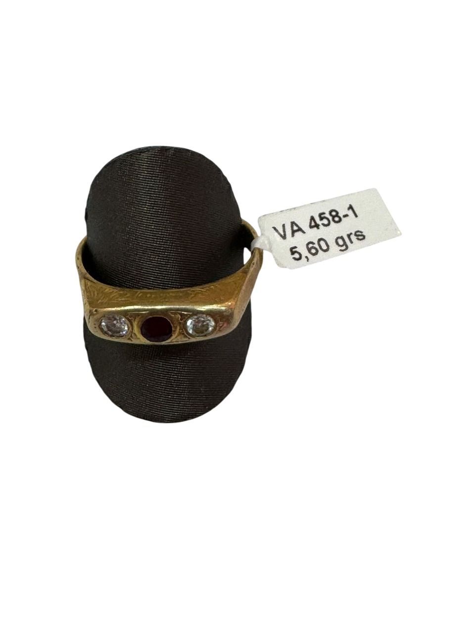 Anillo de oro con una piedra roja y dos piedras transparentes sobre un soporte de anillo negro, con etiqueta de precio.
