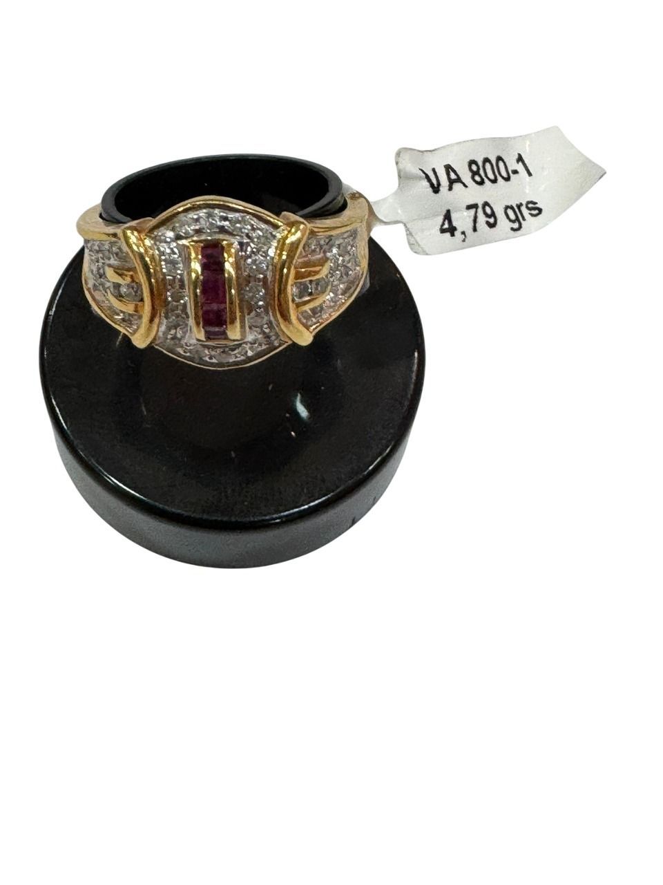 Anillo de oro con diamantes y una piedra preciosa central roja sobre un soporte negro, con una etiqueta de precio.