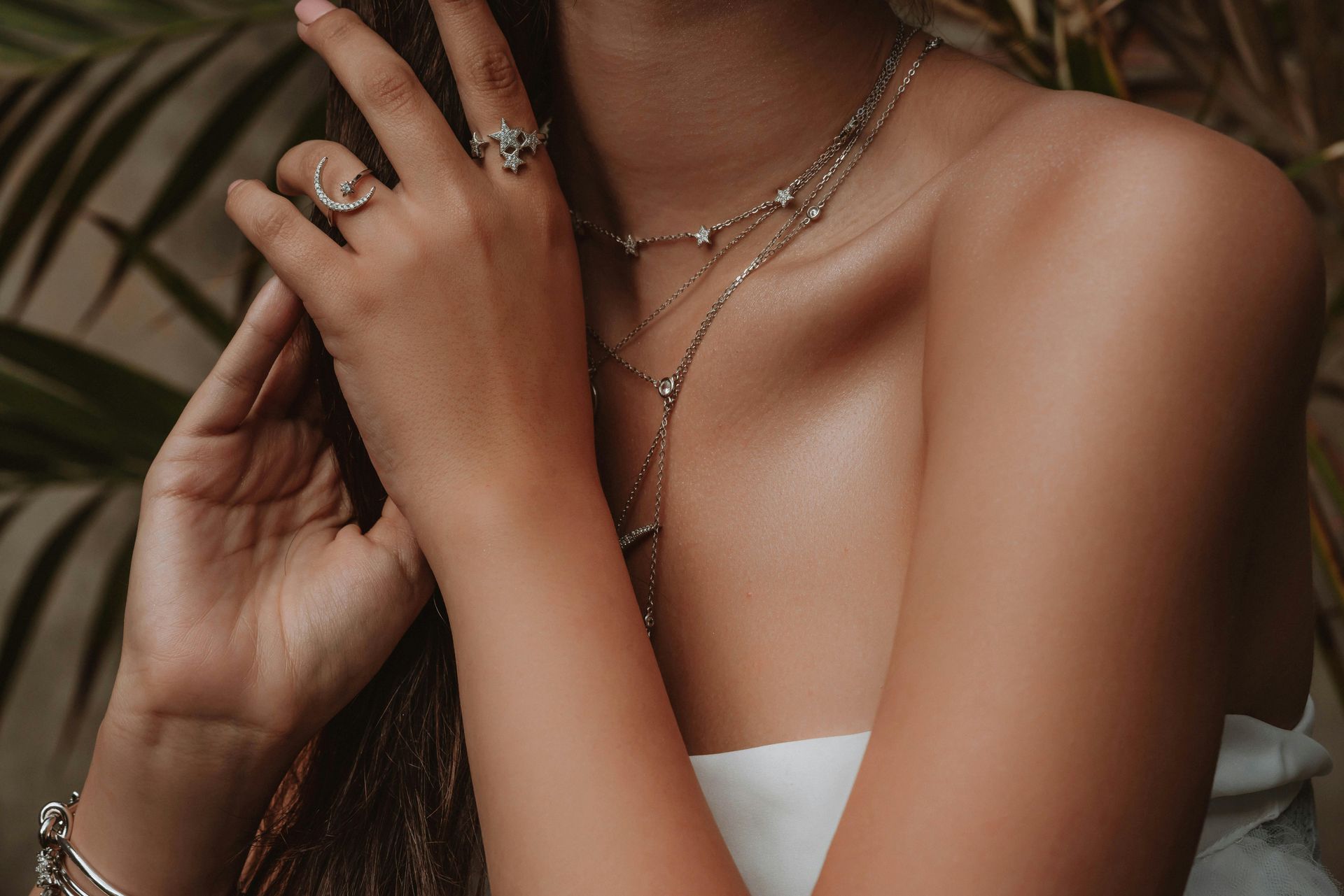 Brazo de mujer con joyas: anillos y collares en capas. Piel bronceada, fondo natural, sujetando el cabello.