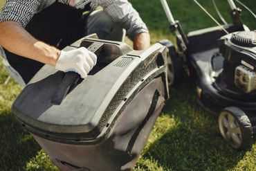 Une personne portant des gants blancs fixe un sac de ramassage d'herbe noir à une tondeuse à gazon sur de l'herbe verte.