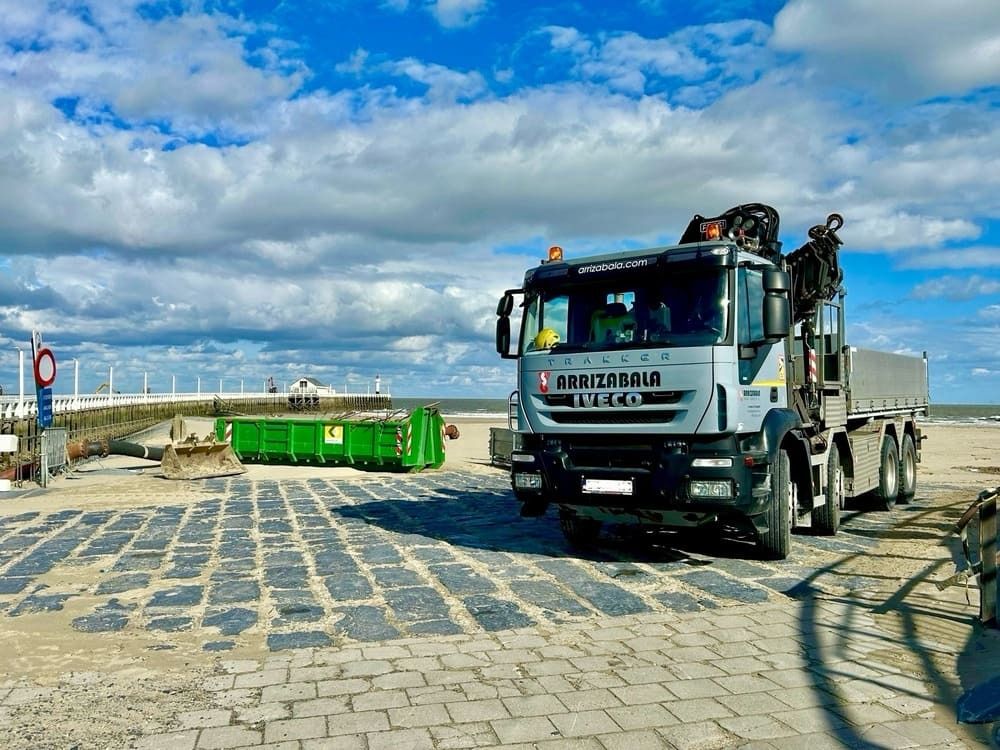 Un camión grande está estacionado en un camino de ladrillos al lado de un contenedor de basura verde.