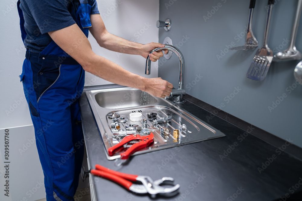 Eine Person in blauem Arbeitsoverall repariert einen Küchenspülenhahn; Werkzeuge liegen auf der Arbeitsfläche.