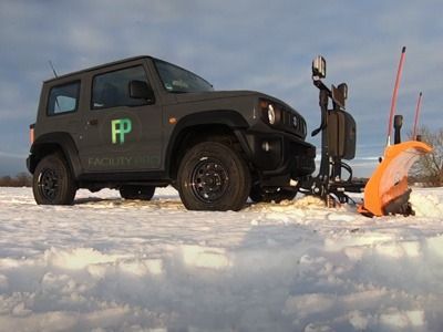 Ein grauer Suzuki Jimny Schneepflug, geparkt auf einem schneebedeckten Feld unter bewölktem Himmel.
